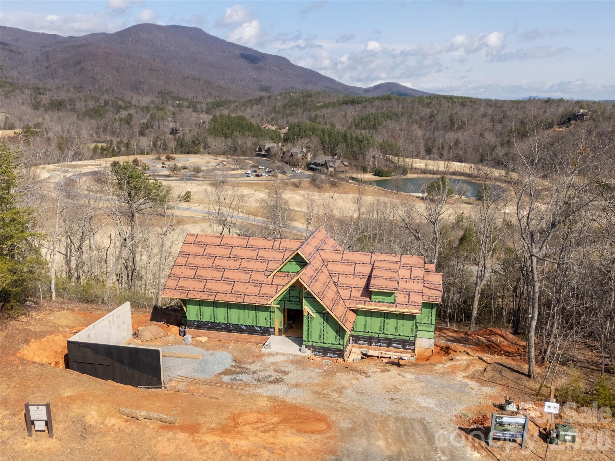 House under construction amid leafless woods, with a pond and mountain ridges beyond.
