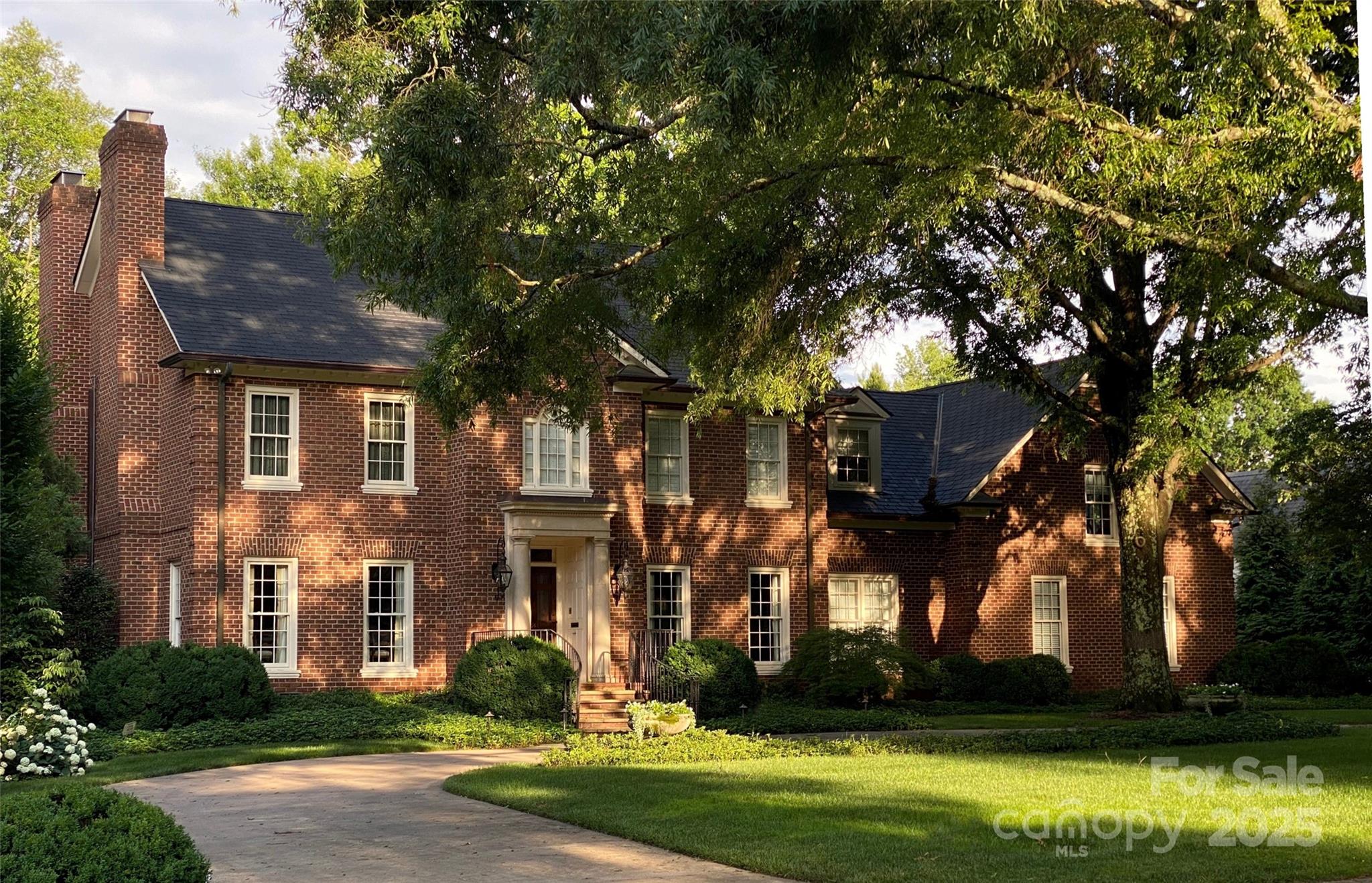 Two-story brick house with white-trimmed windows, a large shade tree, and a curved driveway.