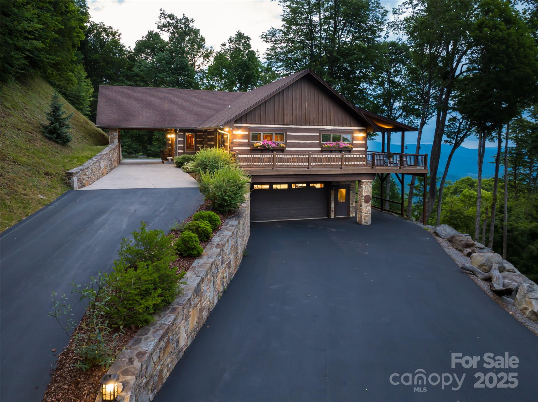 Mountain home with a wraparound deck, stone retaining walls, and a wooded mountain view at dusk.