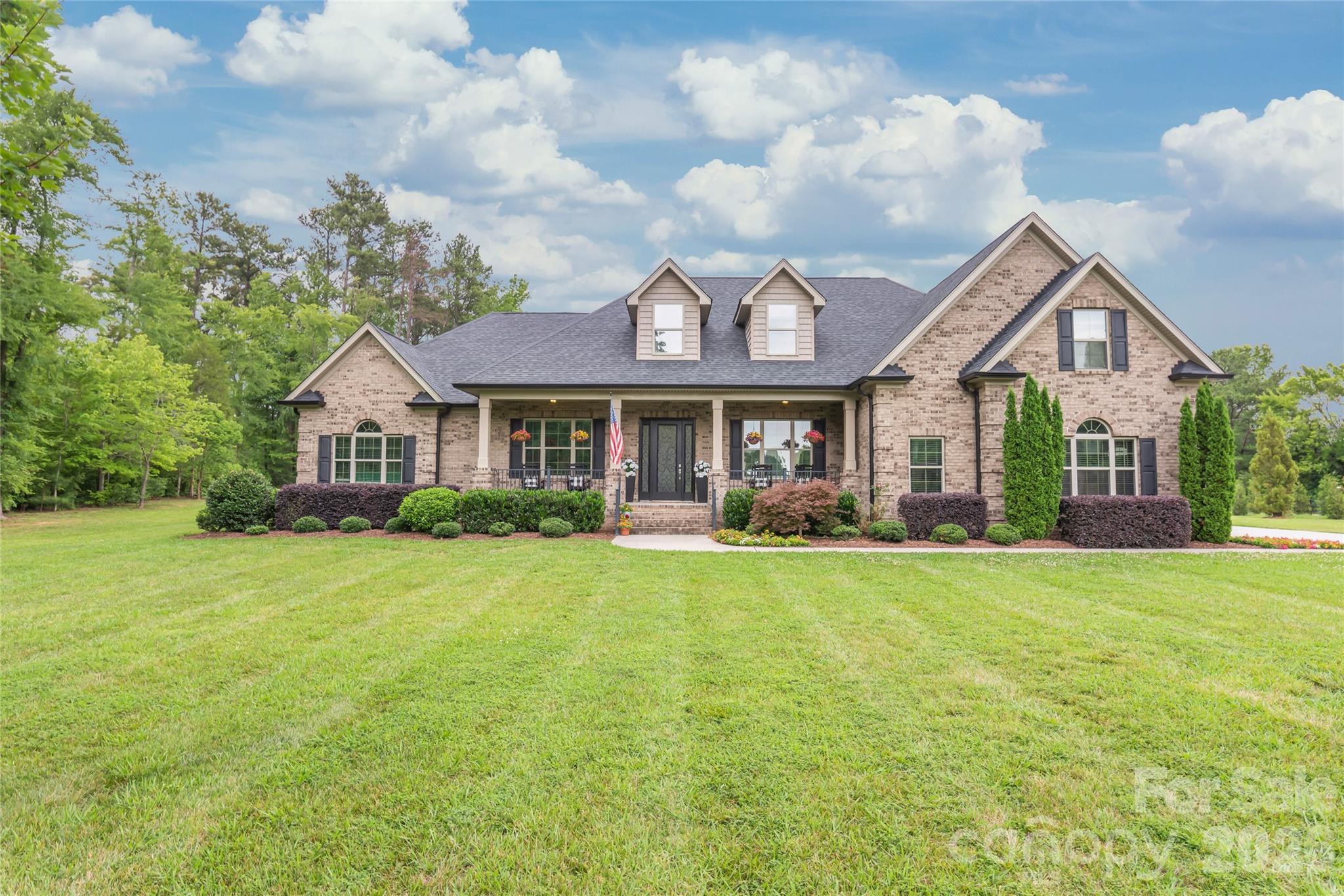 Brick home with a covered porch, roof dormers, landscaped shrubs, and a broad green lawn.
