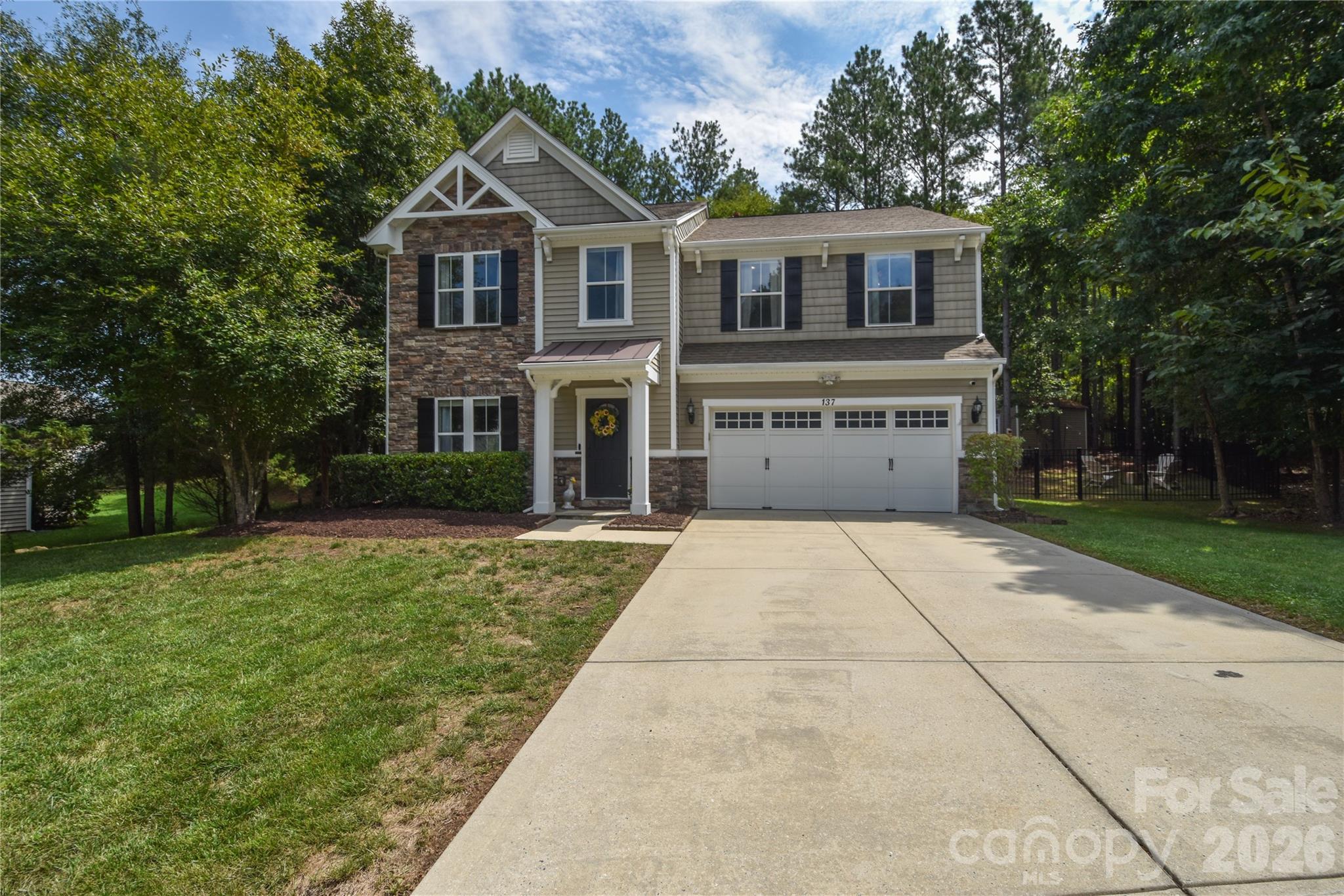 Two-story beige house with stone accents, a double garage, and a wide driveway among mature trees.