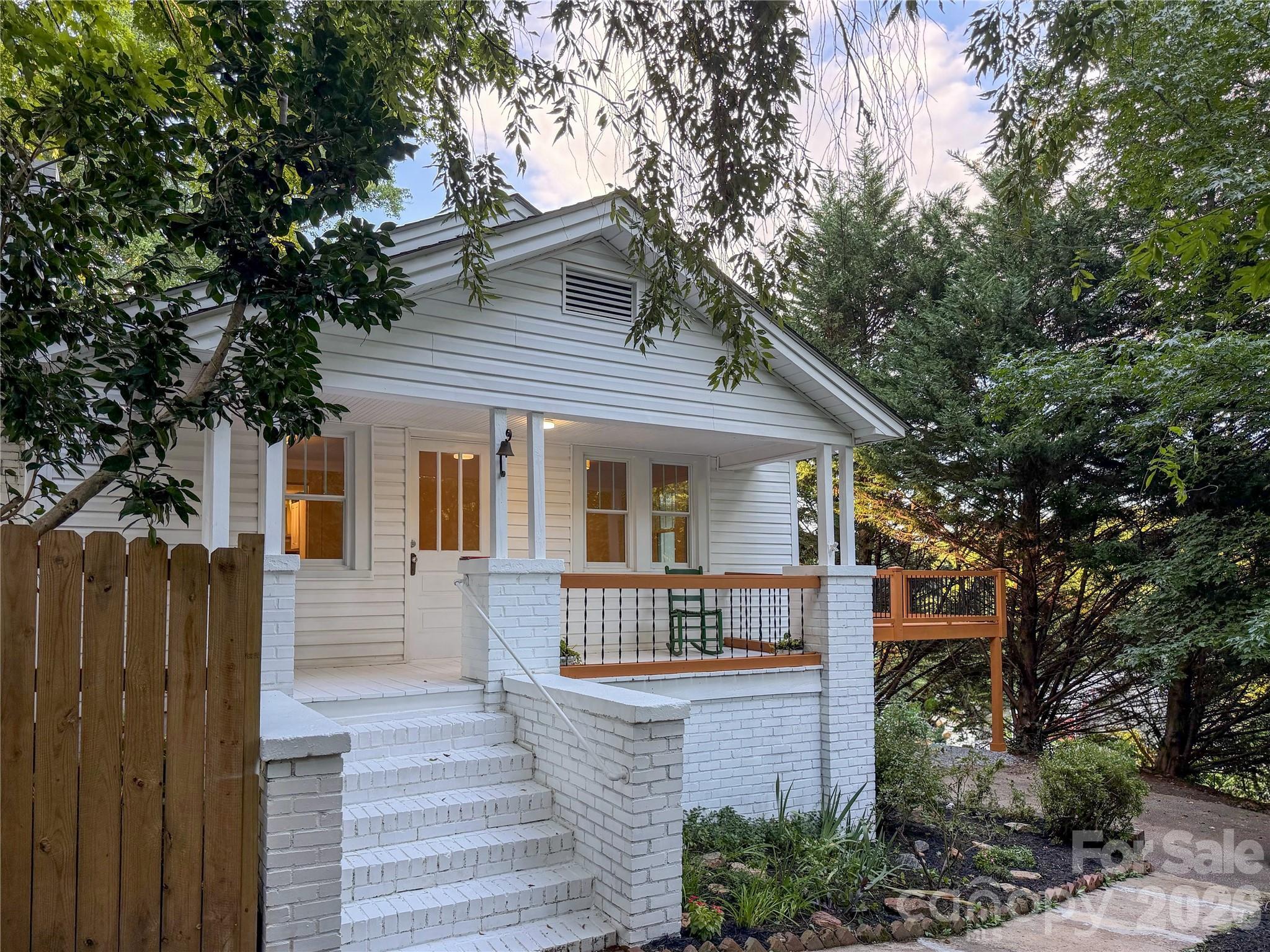 White-sided house with a covered porch, brick steps, and a wooden deck among mature trees.