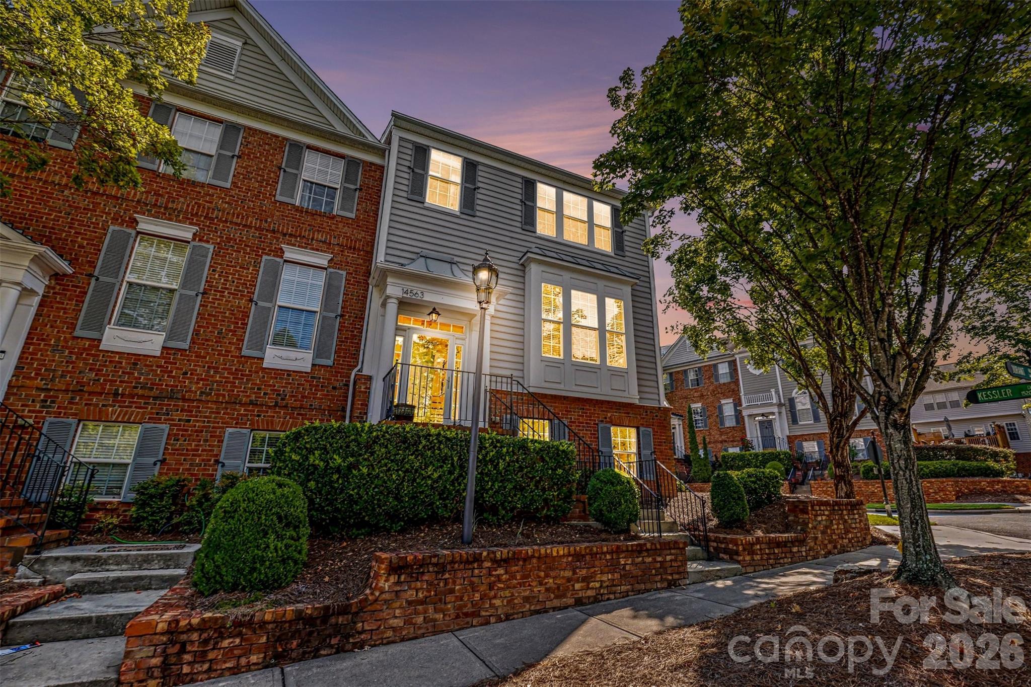 Townhouse exterior at dusk with illuminated windows, brick façade, landscaped entry, and mature trees.