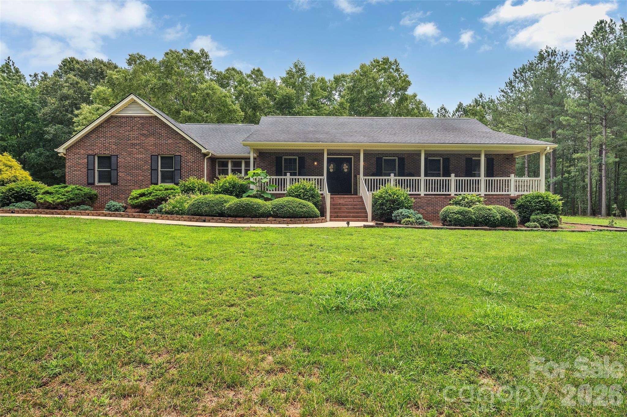 Red brick house with a long covered porch, landscaped shrubs, and a broad front lawn.