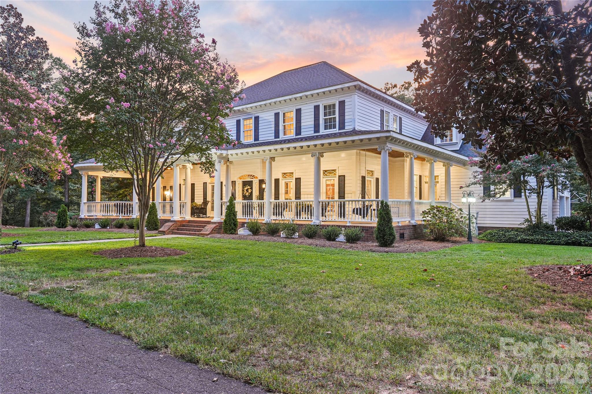 Two-story white house with a columned wraparound porch, glowing lights, and flowering trees at dusk.